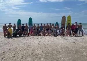 A large group of students posing at the beach with surfboards and other gear for beach activities.